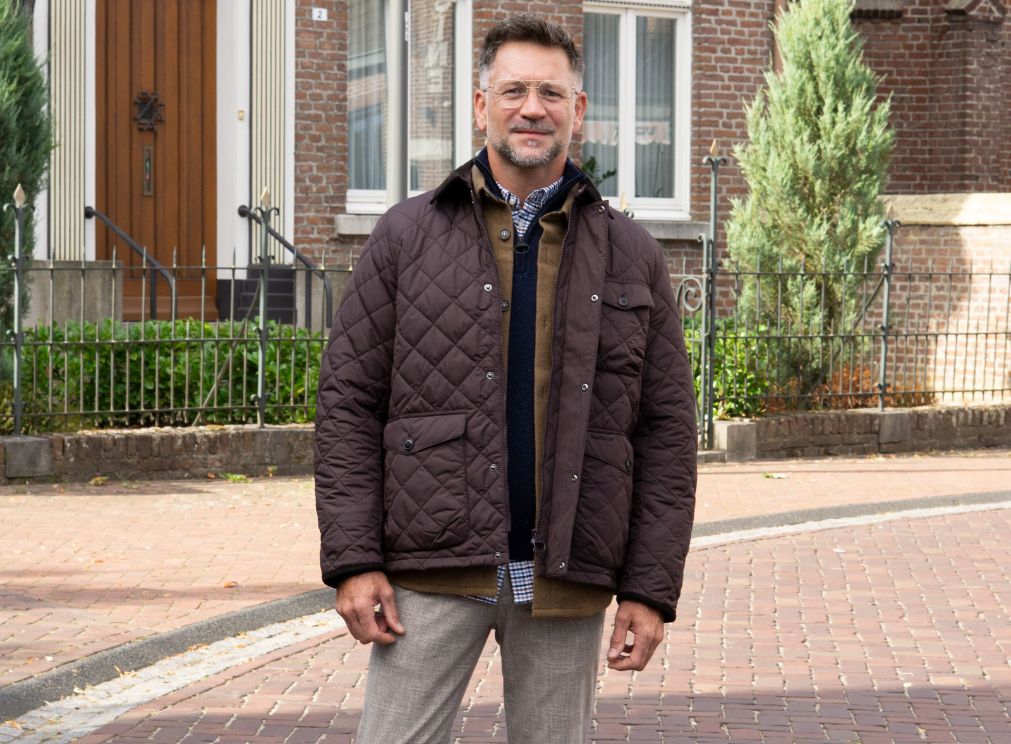 Herenmodel in een donkerbruine gewatteerde jas over een bruin vest, donkerblauwe trui en geruit overhemd, gecombineerd met een lichtgrijze broek.