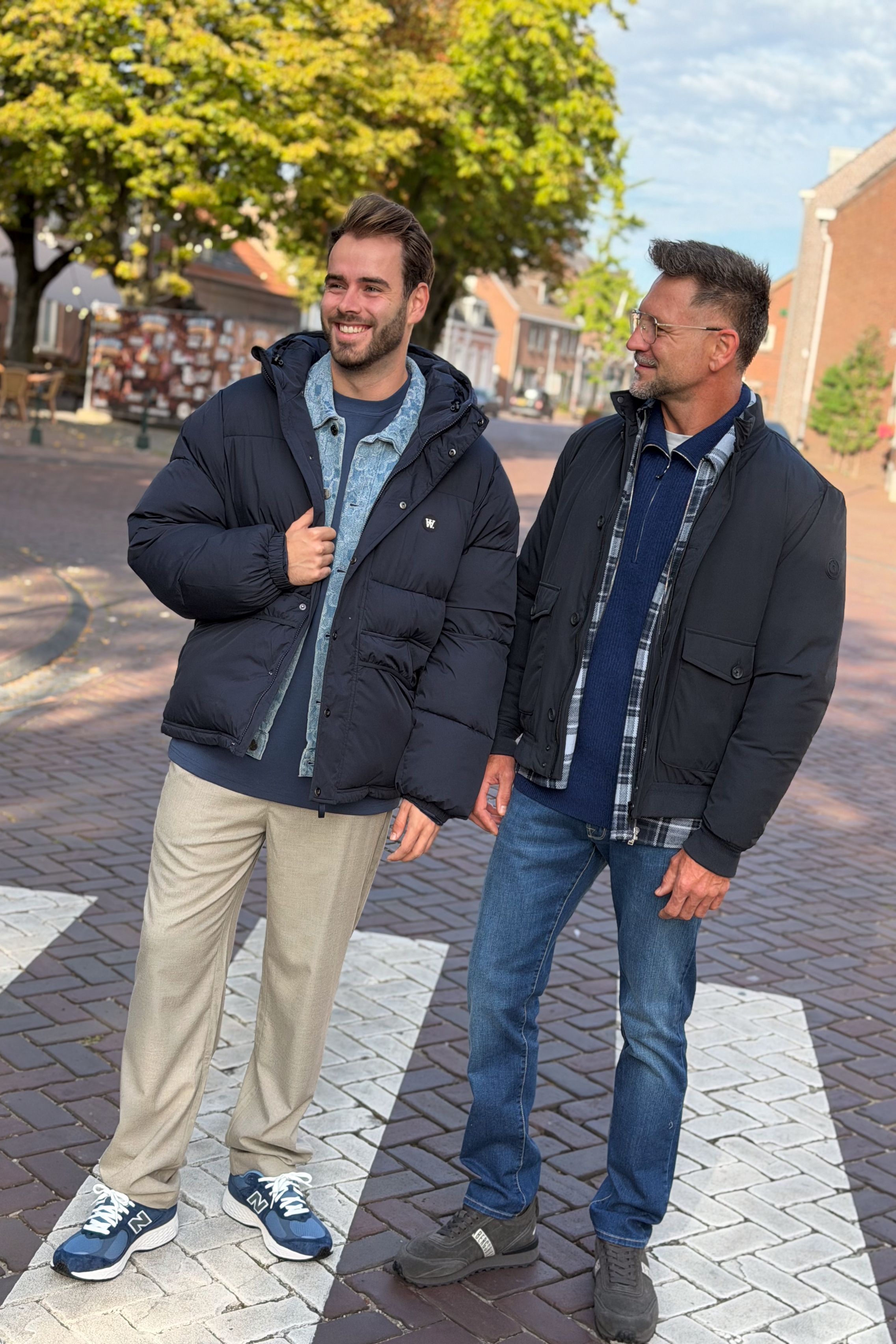 Twee heren in casual najaarslooks: één in een donkerblauwe pufferjas over een denim overshirt met beige broek, de ander in een donkerblauwe jas met geruit overshirt, blauwe trui en jeans.