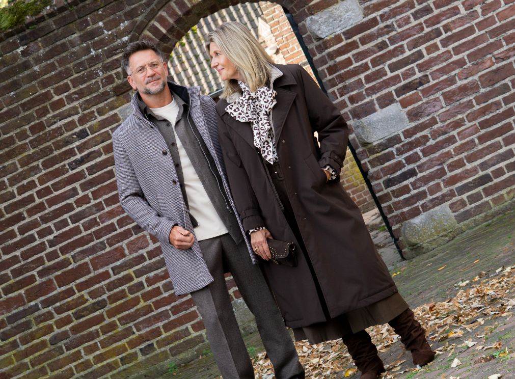 Dames- en herenmodel in stijlvolle najaarsoutfits. Zij draagt een lange donkerbruine jas met een blouse met dierenprint en hoge bruine laarzen. Hij draagt een grijs gemêleerde jas over een bruin vest en lichte trui, gecombineerd met een grijze broek.