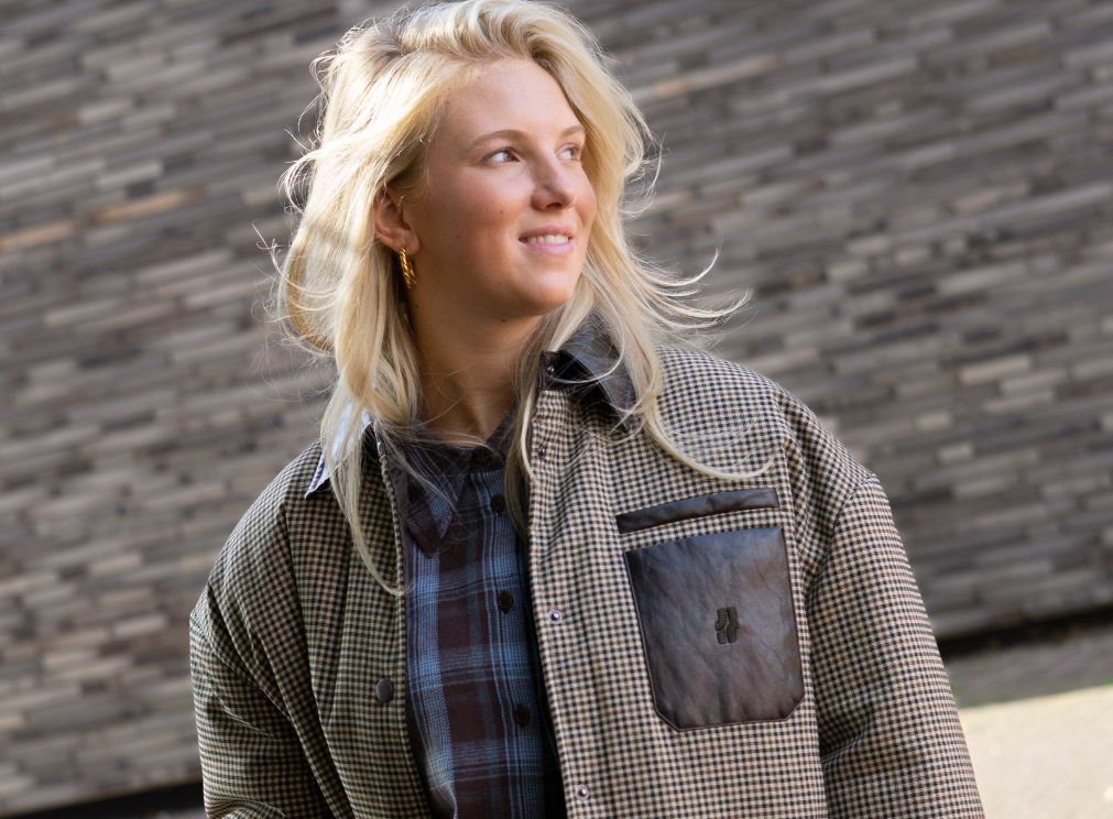 Damesmodel in een geruite jas met bruine leren details, gedragen over een blauw geruite blouse.